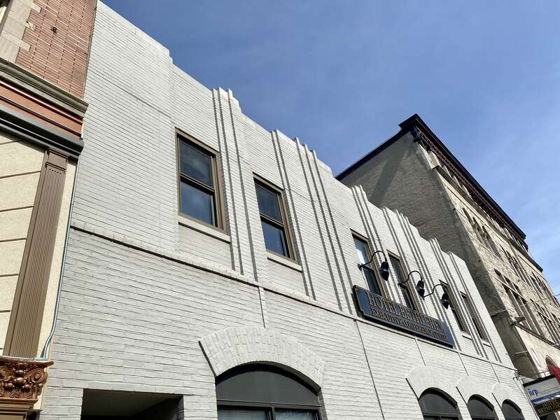 Built in the early 20th Century, this Art Deco-style building features a painted brick exterior, pilasters that stair-step in and out of the facade with vertical lines of single bricks in the middle of each pilaster except at the ends, recessed