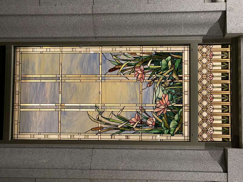 A stained glass window from the Orem Utah Temple, depicting bullrushes and cherry blossoms, next to water elements, with a blue sky with clouds. A bronze rectangular frame frames it and has stone masonry surrounding the window.
