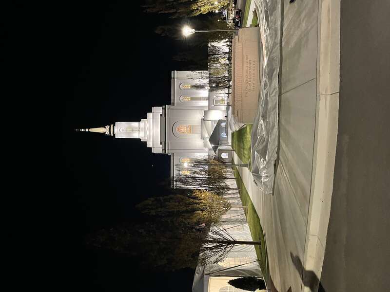 A picture of the Orem Utah Temple at night. Garden elements can be seen in the foreground, plastic wrapping shows underneath a sign that says: "The Church of Jesus Christ of Latter Day Saints, Orem Utah Temple."