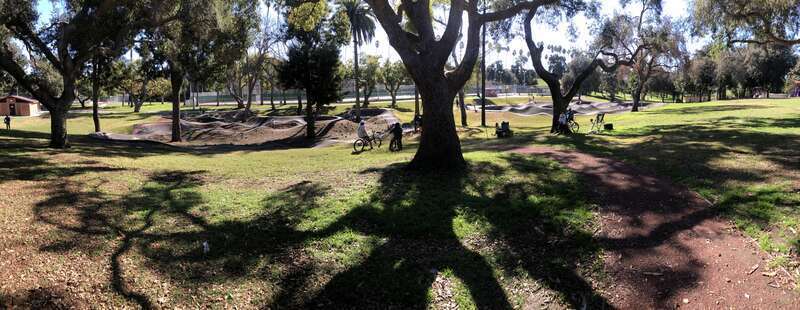 This is a midday photograph of the Inglewood Pumptrack from the north, facing south toward the bike facility. The Pumptrack's two tracks are visible in the picture. Just left of center is a looping track for riders to travel on BMX or mountain bikes.