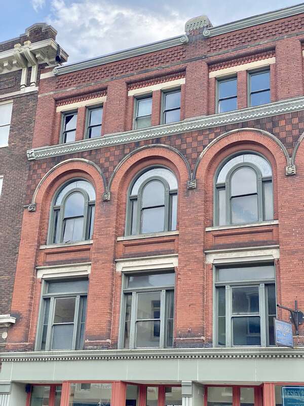 Built in 1895, this Romanesque Revival-style building features a red brick exterior, arched windows on the third floor, stone trim, decorative patterned brickwork, one-over-one double-hung windows, and a modified first floor retail shopfront.  The