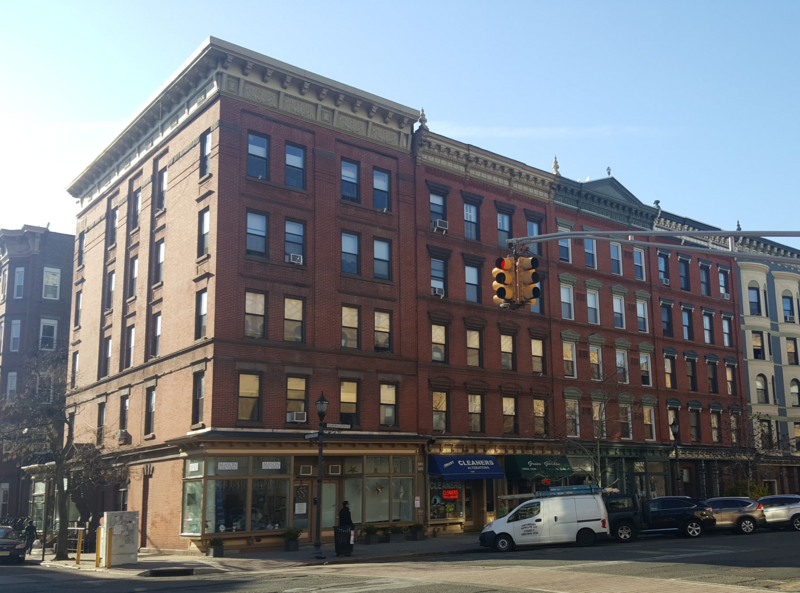 The buildings on the northwest corner of 12th and Washington Streets in Hoboken, New Jersey.  Picture taken from the southeast corner of the intersection, looking west-by-northwest.At the bottom left, just to the right of the tree and the pedestrian,