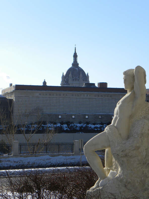 Minnesota History Center and the St. Paul Cathedral in the background with "Earthbound, Made Captive, Yet Deserving Freedom More" a 1956 marble sculpture by John Karl Daniels in the foreground on the Minnesota State Capitol Mall.