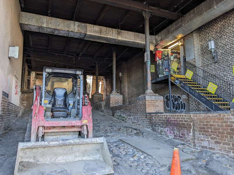A ramp down to River Street underneath the former Savannah Cotton Exchange, with stairs to a shop side entrance and a parked skid steer.