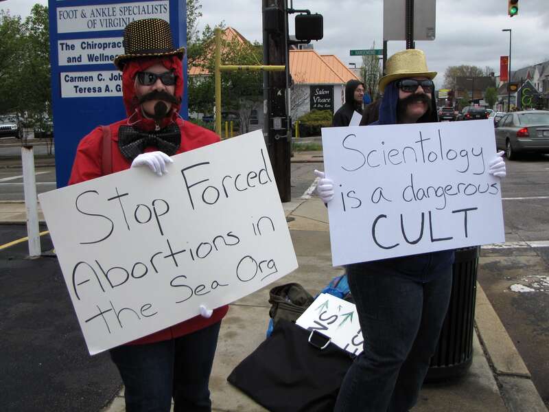 Arcadia and HT, from the Washington DC Anonymous cell, at the Church of Scientology mission in Richmond, Virginia.  Arcadia holds a sign saying, "Stop Forced Abortions in the Sea Org", and HT holds one that says "Scientology is a dangerous CULT".