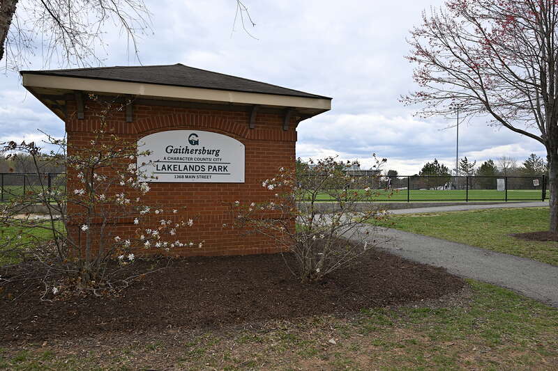 The sign for Lakelands Park with flowering bushes by it and playing fields behind it. This park is run by the city of Gaithersburg, Maryland. 1368 Main Street, Gaithersburg, Maryland 20878.