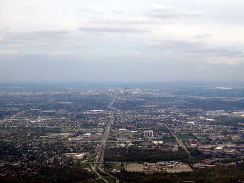 Aerial photograph of Dearborn, Michigan with downtown Detroit and Windsor, Canada in background. Ford World Headquarters is partially visible on far right, other Ford buildings visible throughout the image.