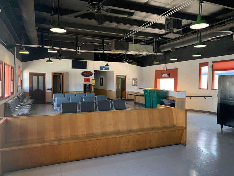 The interior waiting area of the Amtrak station in Grand Junction, Colorado. Taken during daylight hours standing near the street side entrance looking toward the trackside entrance. Visible amenities include benches, chairs, trash bins, payphone,