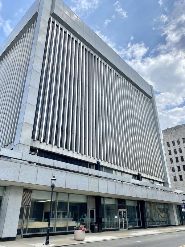 Built in 1969, this Modern International-style skyscraper features a stone panel and metal-clad exterior with a one-story podium featuring a large parapet and dark stone-clad columns, a plaza atop the roof of the podium with planters and flagpoles,