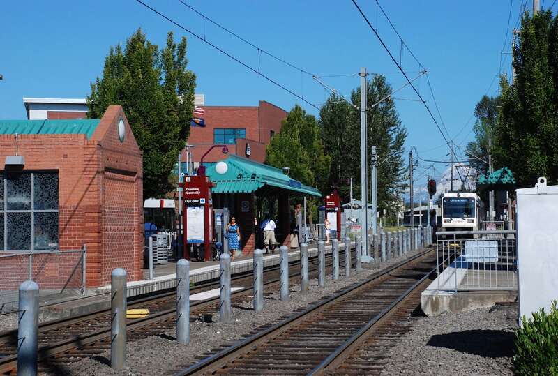 The MAX light rail station at TriMet's Gresham Central Transit Center, viewed from the west.  An outbound train is pulling away from the platform (this is the rear of the train).  Mount Hood is visible in the distance.The building at left was