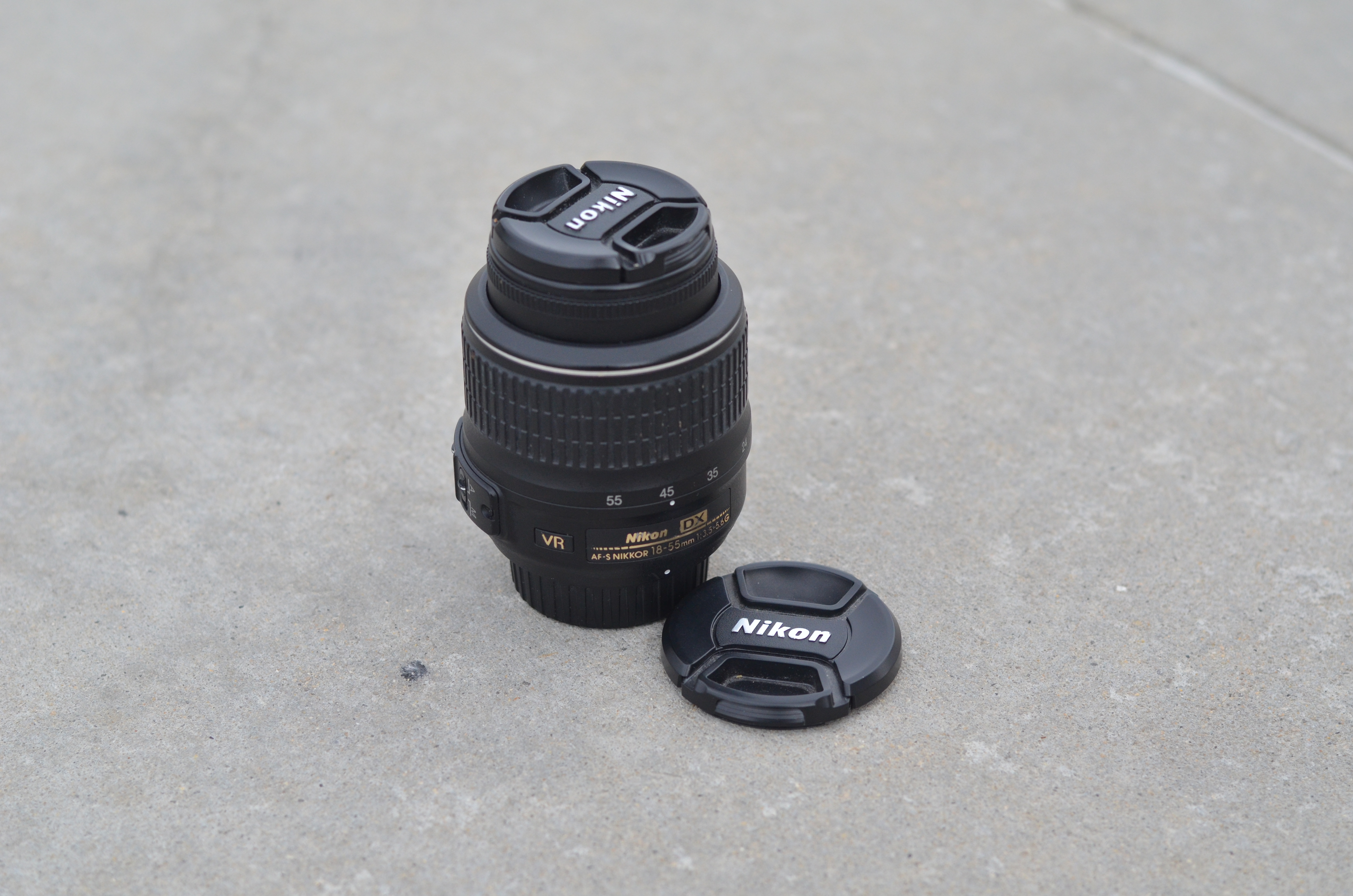 A Nikon DX AF-S 18-55mm VR F-Mount lens, adjusted to 45mm focal length, is pictured next to a Nikon brand lens cap identical to that covering the lens to the right. The picture is shot outdoors at an angle from above, on a concrete pavement surface