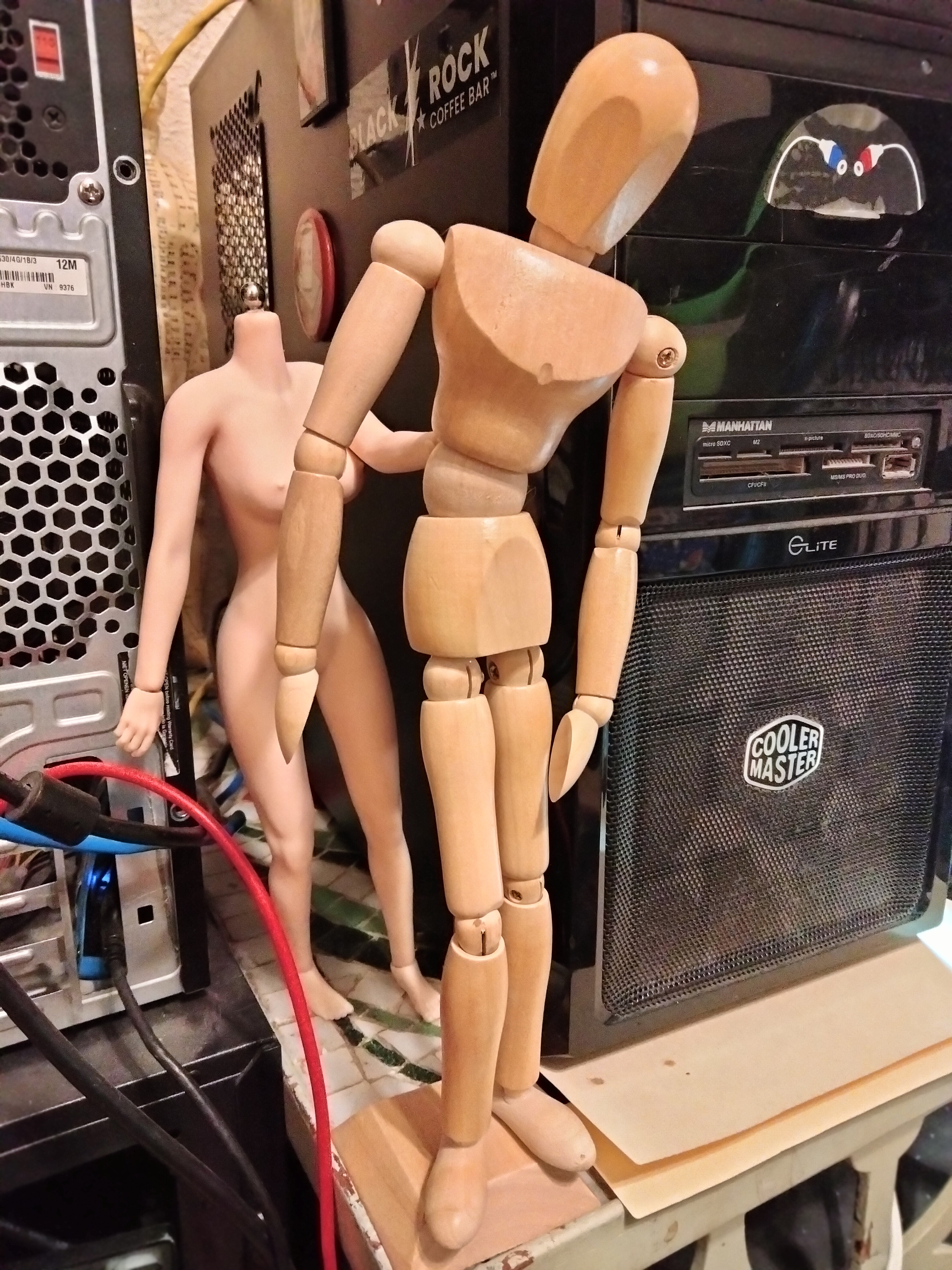 Two 30.48 cm lay figures stand on a table between with two computers with black cases; the wooden nonbinary figure is of the abstract sor whereas the female is seamless, anatomically correct, and flesh-colored pale.